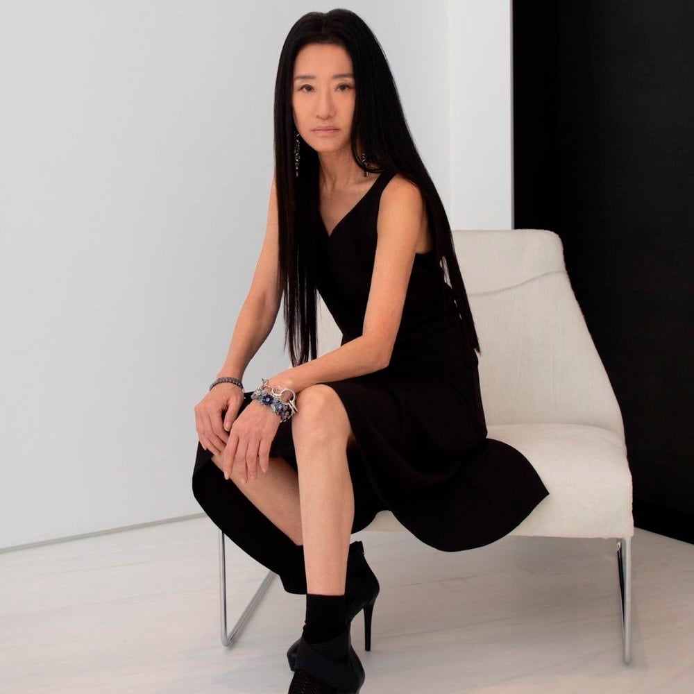 A woman with long straight black hair sits on a white modern chair in a minimalistic room. She wears a sleeveless black dress, black ankle boots, and silver jewelry, and looks directly at the camera with a calm expression.