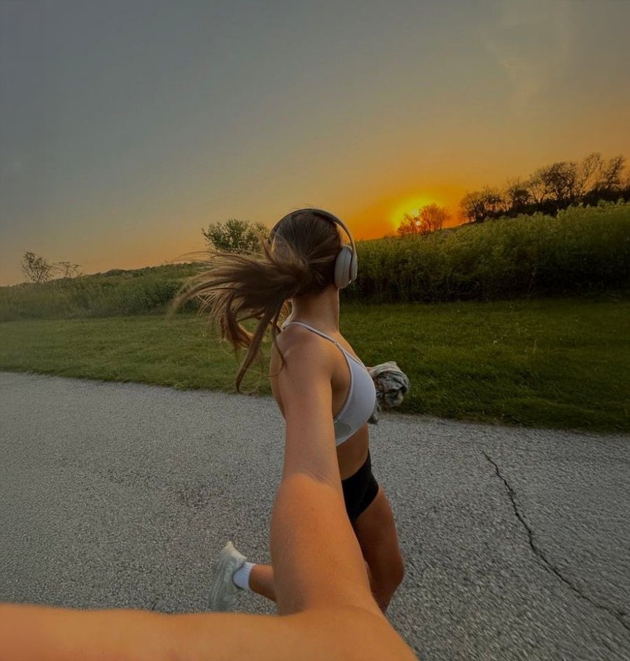 A woman in athletic wear runs on a paved road at sunset, wearing headphones and holding someone’s hand. Her hair flows behind her, and greenery lines the road under a colorful sky.