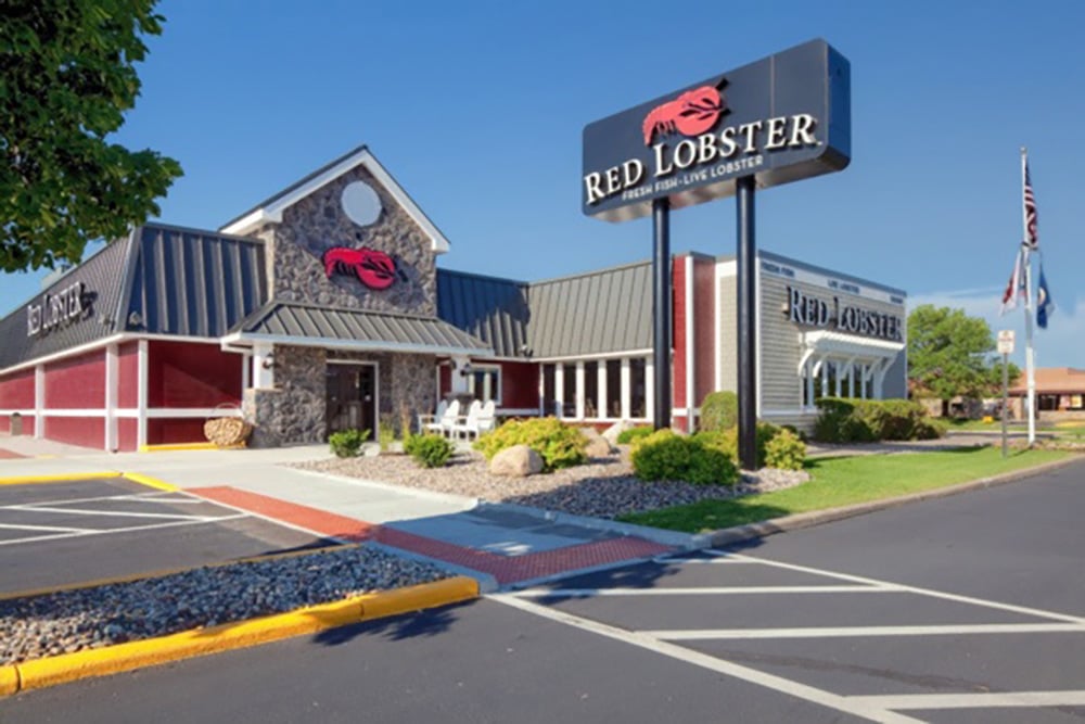 A Red Lobster restaurant building with a large sign in front, featuring the red lobster logo. The exterior has red siding, stone accents, and a neatly landscaped parking lot under a clear sky.