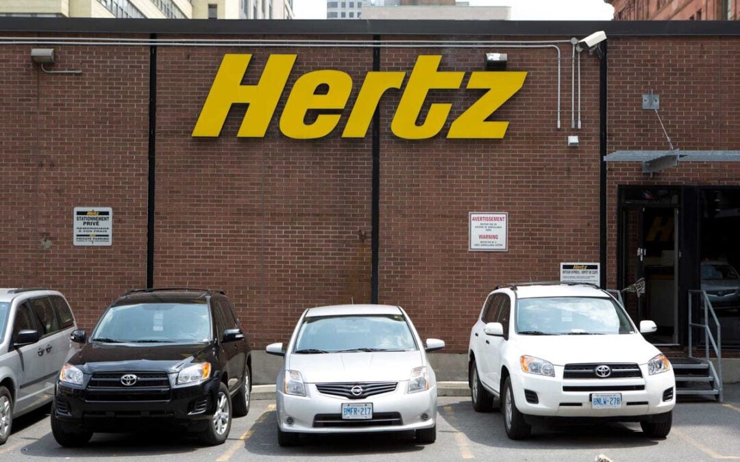 Three cars are parked in front of a brick building with a large yellow "Hertz" sign, indicating a car rental location. The building has a door and several security and parking signs.