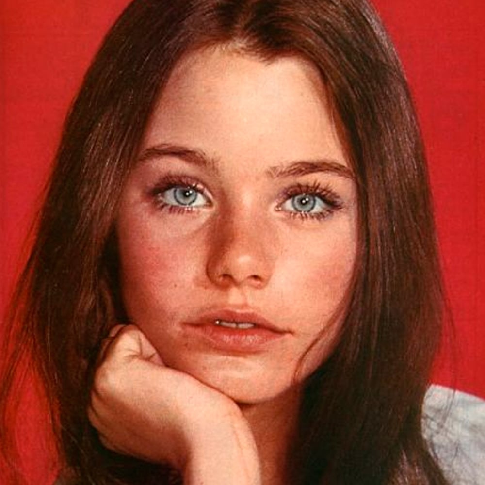 A young woman with long brown hair and blue eyes rests her chin on her hand, gazing forward. She has a natural look with minimal makeup and is posed against a solid red background.