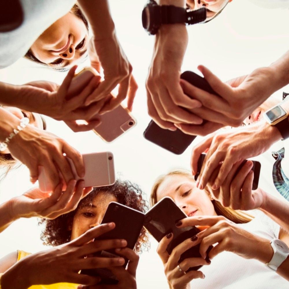 A group of people stand in a circle, holding and using smartphones. The photo is taken from below, capturing their hands and faces as they look down at their screens.