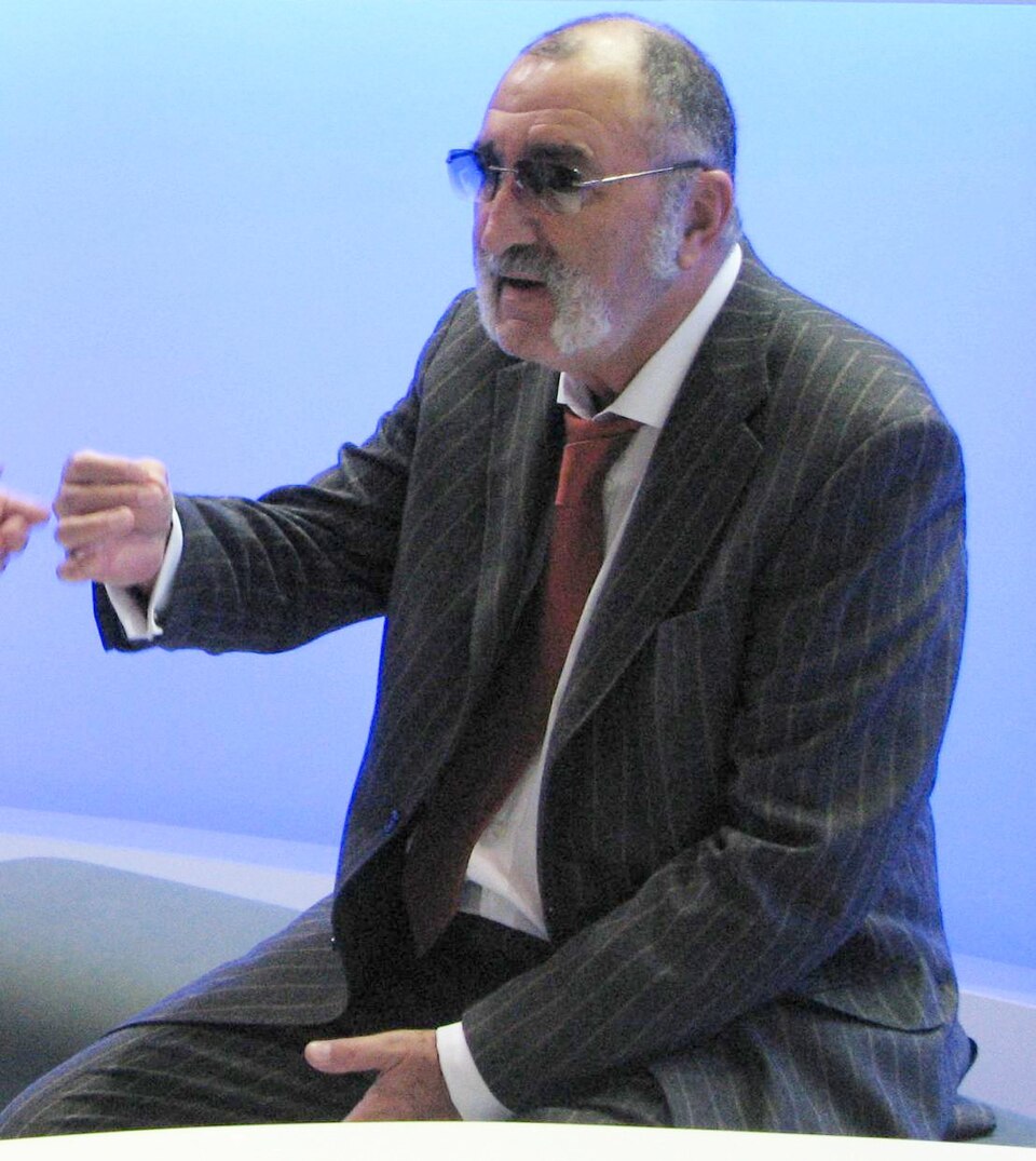 A man with a gray beard and mustache, wearing a pinstripe suit, red tie, and sunglasses, sits gesturing with his right hand against a blue background.