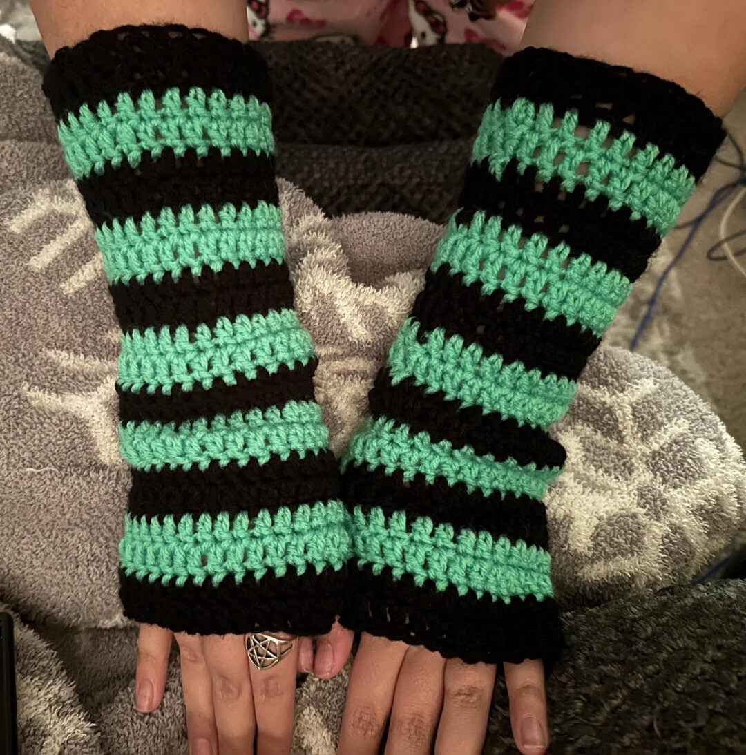 Two hands wearing crocheted fingerless gloves with horizontal stripes in black and mint green, resting on a patterned grey and white blanket.
