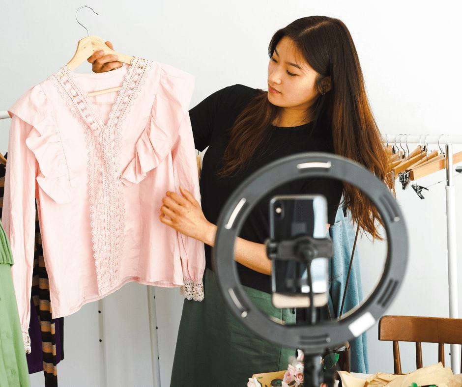 A woman stands behind a ring light and smartphone, holding up a pink ruffled blouse on a hanger, likely showcasing it for an online video or livestream in a clothing store or boutique.