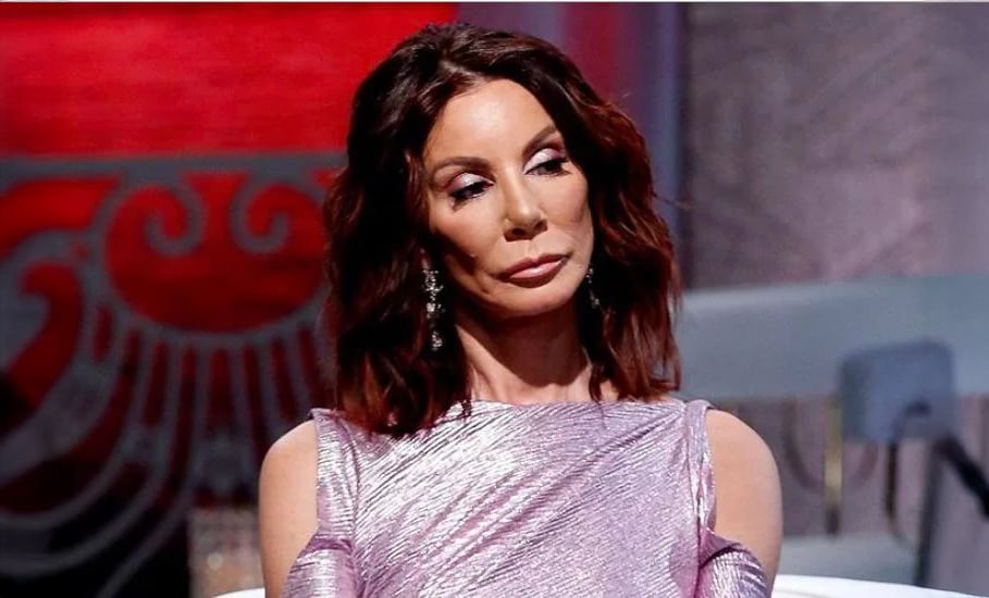 A woman with wavy, shoulder-length brown hair sits in a shiny lavender dress, looking to the side with a neutral expression. The background features a red and gray decorative pattern.