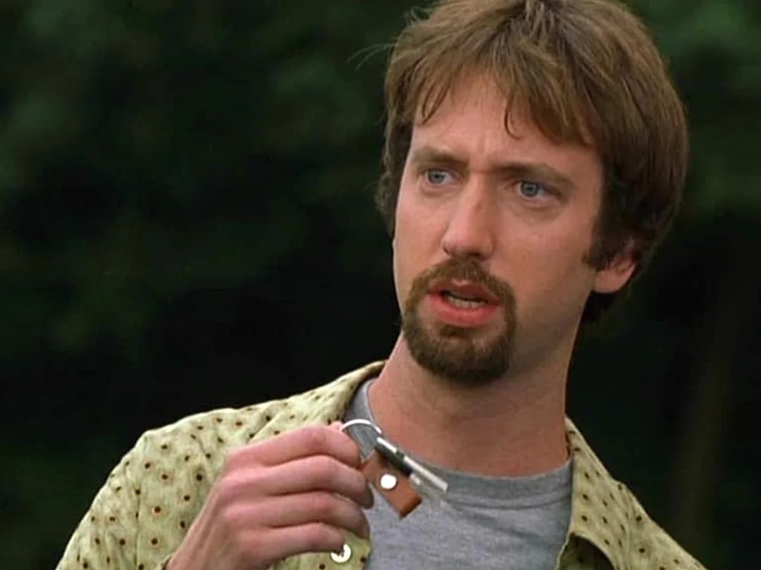 A man with light skin, brown hair, and a goatee holds a small object with a keychain, looking surprised. He wears a light patterned shirt over a gray t-shirt. The background is blurred greenery.