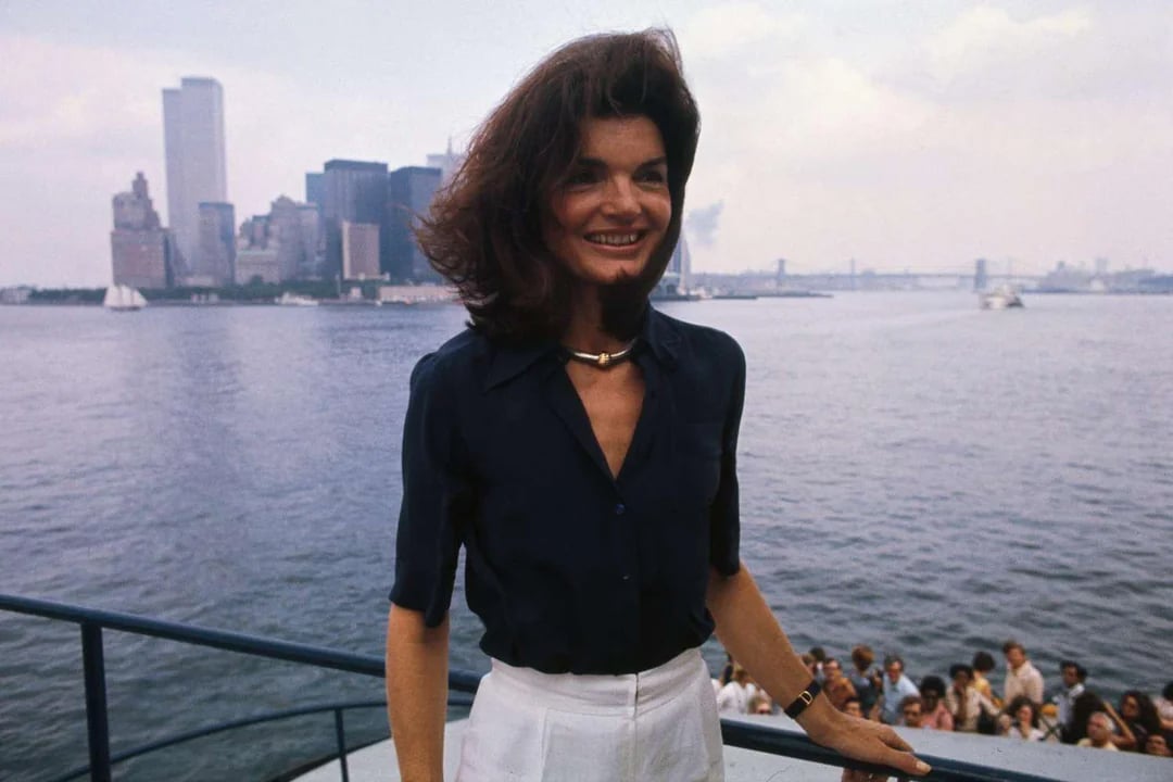 A woman with dark hair in a navy blouse and white pants stands on a boat with a city skyline and water in the background; a crowd is gathered below on the deck.