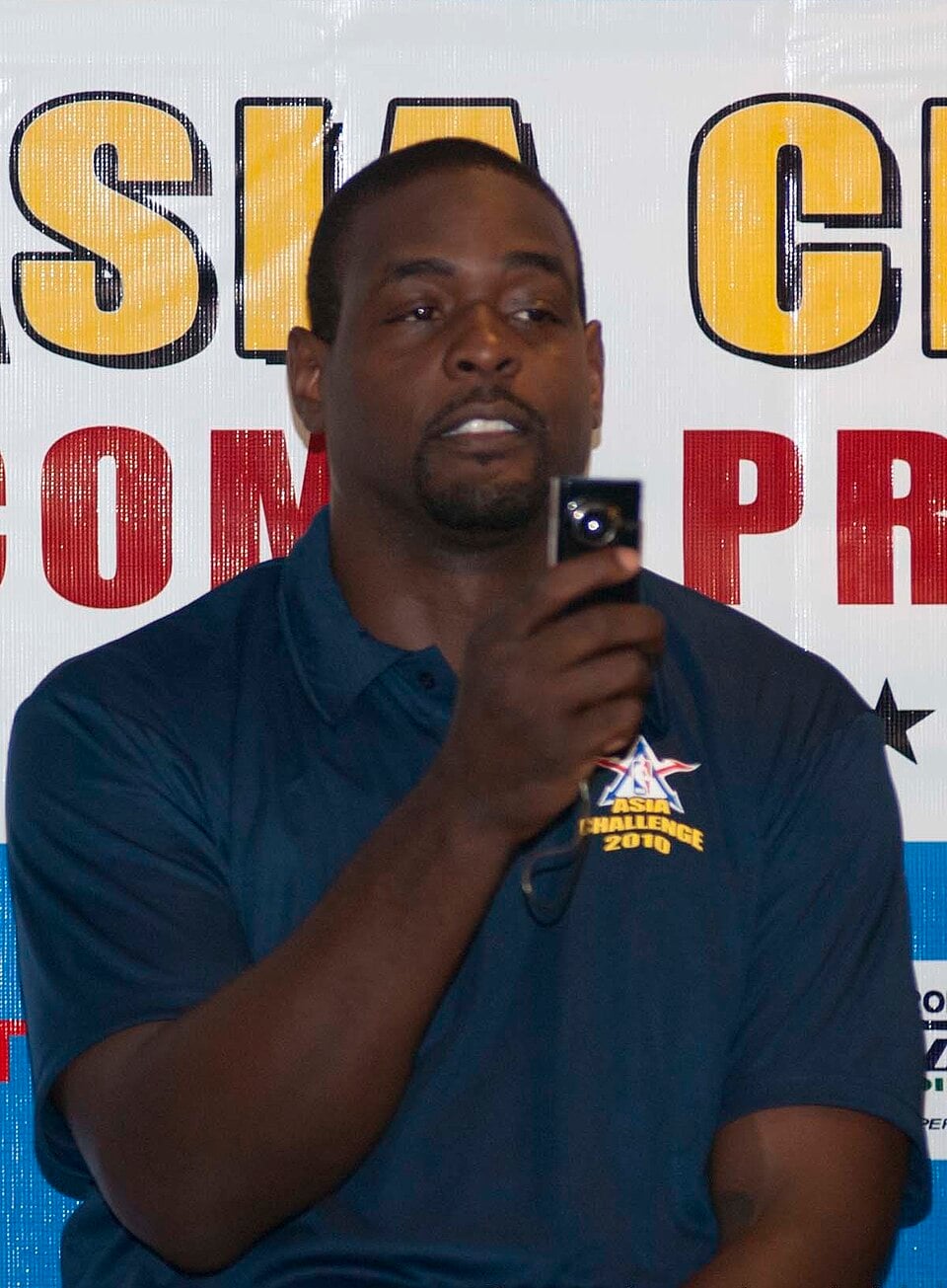 A man in a navy blue polo shirt holds up a small camera while speaking in front of a colorful, partially visible sign with large yellow and red letters.