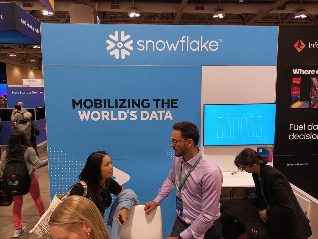 Three people are talking at a trade show booth with a blue display that reads “snowflake” and “Mobilizing the World’s Data.” A screen and informational materials are visible on the booth.
