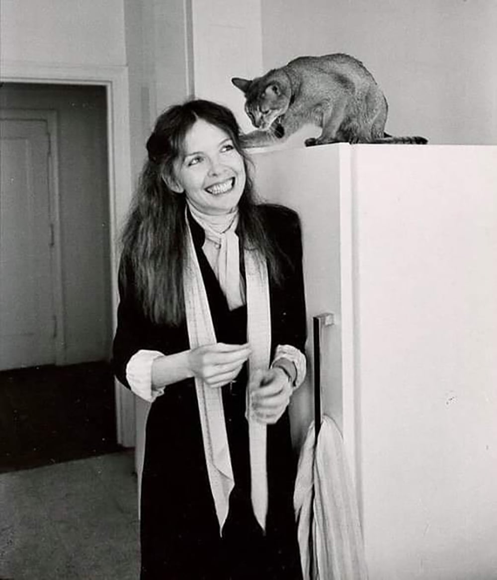 A woman with long hair smiles while standing next to a refrigerator. A cat sits on top of the fridge, looking down at her. The scene is indoors, and the image is in black and white.