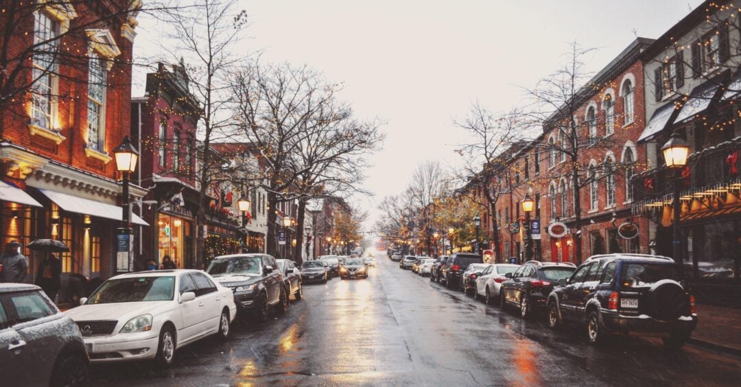 A city street lined with parked cars and brick buildings, decorated with holiday lights. Bare trees and wet pavement reflect the lights, suggesting wintertime and recent rain. People walk on the sidewalks under a cloudy sky.