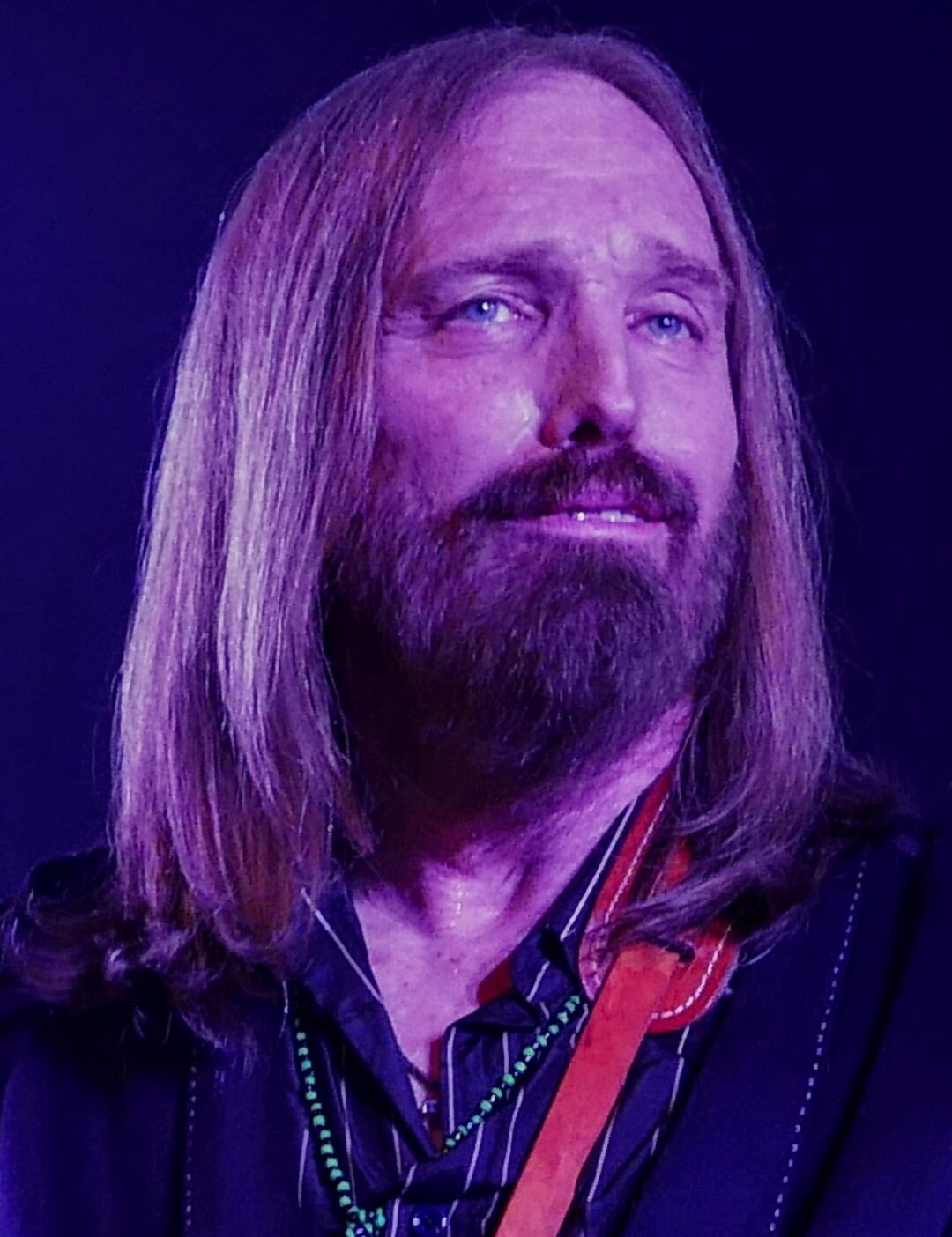 A man with long, straight, light brown hair and a beard wears a dark shirt and beaded necklace, looking slightly upward and smiling softly. The background is dark purple.