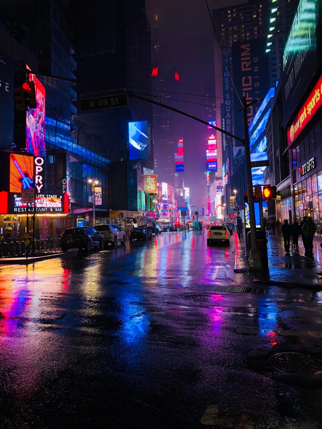 A rainy night in Times Square, New York City, with colorful neon lights reflecting on the wet street. Cars and people are visible along the sidewalks, and bright billboards light up the scene.