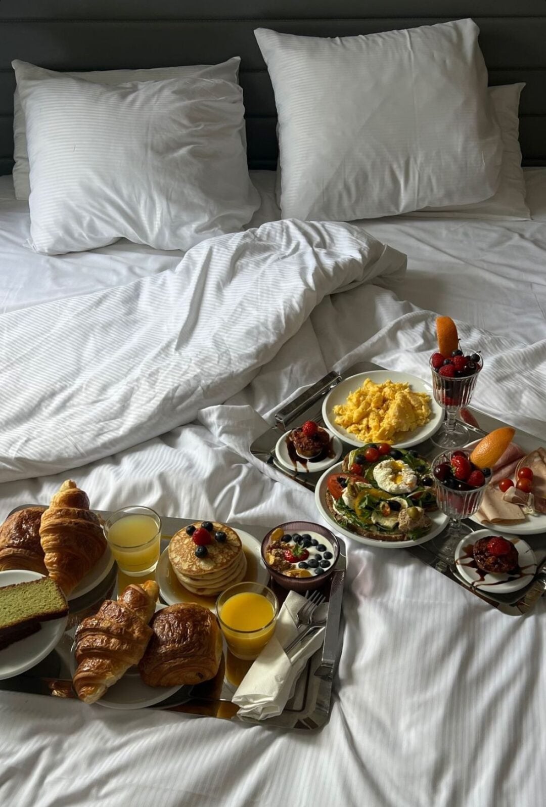 Breakfast trays on a bed with white sheets and pillows. The trays hold croissants, pancakes with berries, juice, eggs, salad, yogurt with fruit, and various pastries and desserts, set for two people.