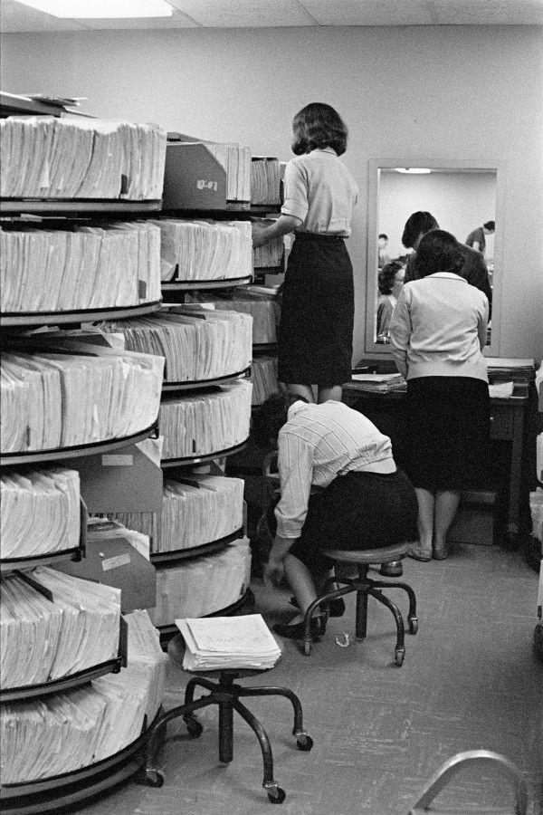 Three women work in a records room filled with rotating shelves of paper files; one stands on a chair to reach a high shelf, another bends down, and the third stands at a desk facing a mirror.