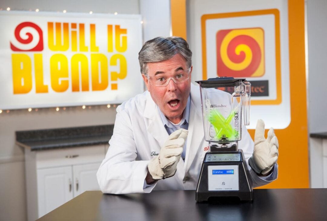 A man in a white lab coat and gloves excitedly poses next to a blender with tennis balls inside. Behind him is a sign that reads "Will It Blend?" in a brightly lit room.