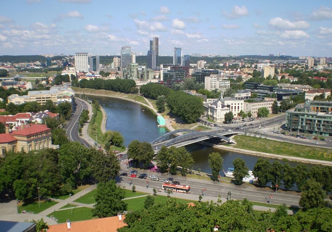 A cityscape with a river curving through the center, modern high-rise buildings in the background, tree-lined streets, bridges, and a mix of historic and contemporary architecture under a partly cloudy sky.