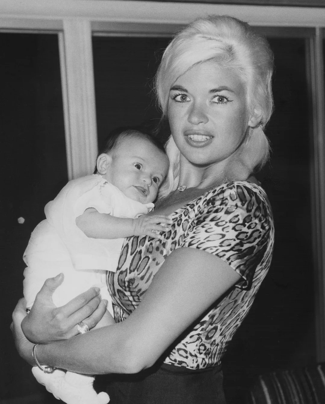 A woman with light hair, wearing a leopard-print top, holds a smiling baby dressed in white in her arms. They are indoors, standing in front of large windows.