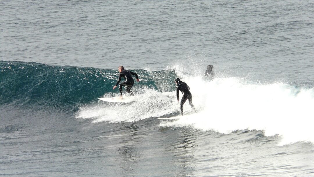Two surfers in wetsuits ride the same wave, with one ahead and the other closely behind. A third surfer is partially visible in the background, waiting in the water. The ocean appears calm except for the breaking wave.