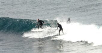 Three surfers in wetsuits ride a wave in the ocean; two are on their boards in the foreground, while a third surfer is further behind, partially obscured by the water&rsquo;s spray.