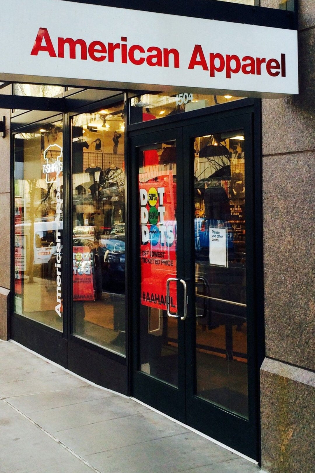 Street view of an American Apparel store entrance with glass doors, sale signs, and clothing displays visible inside. The store’s red and white sign hangs above the door.