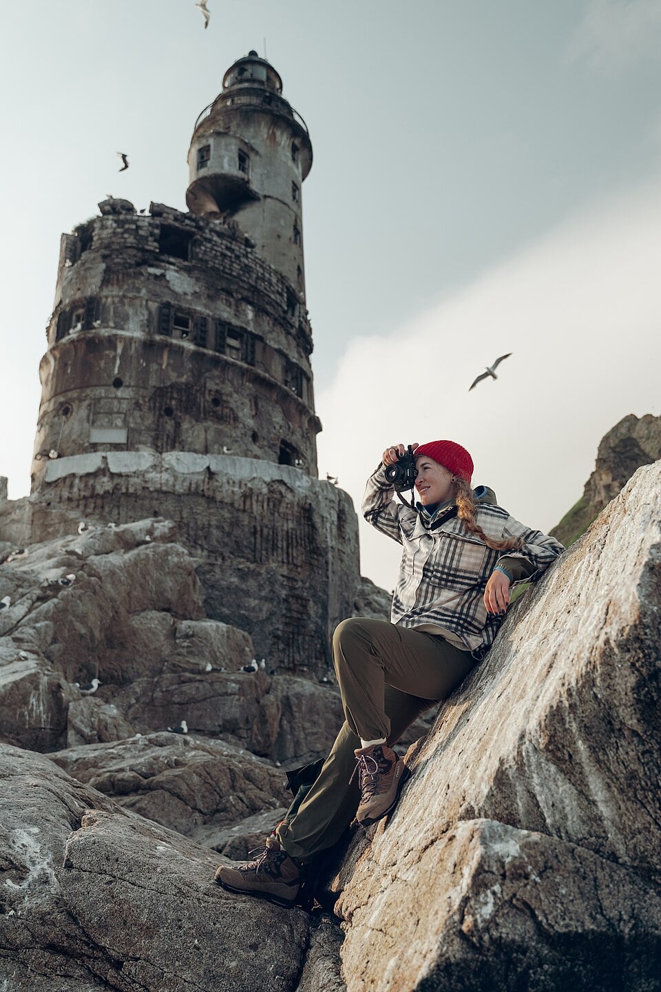 A woman in outdoor clothing and a red beanie sits on rocky terrain, looking through binoculars near a weathered lighthouse with birds flying overhead.