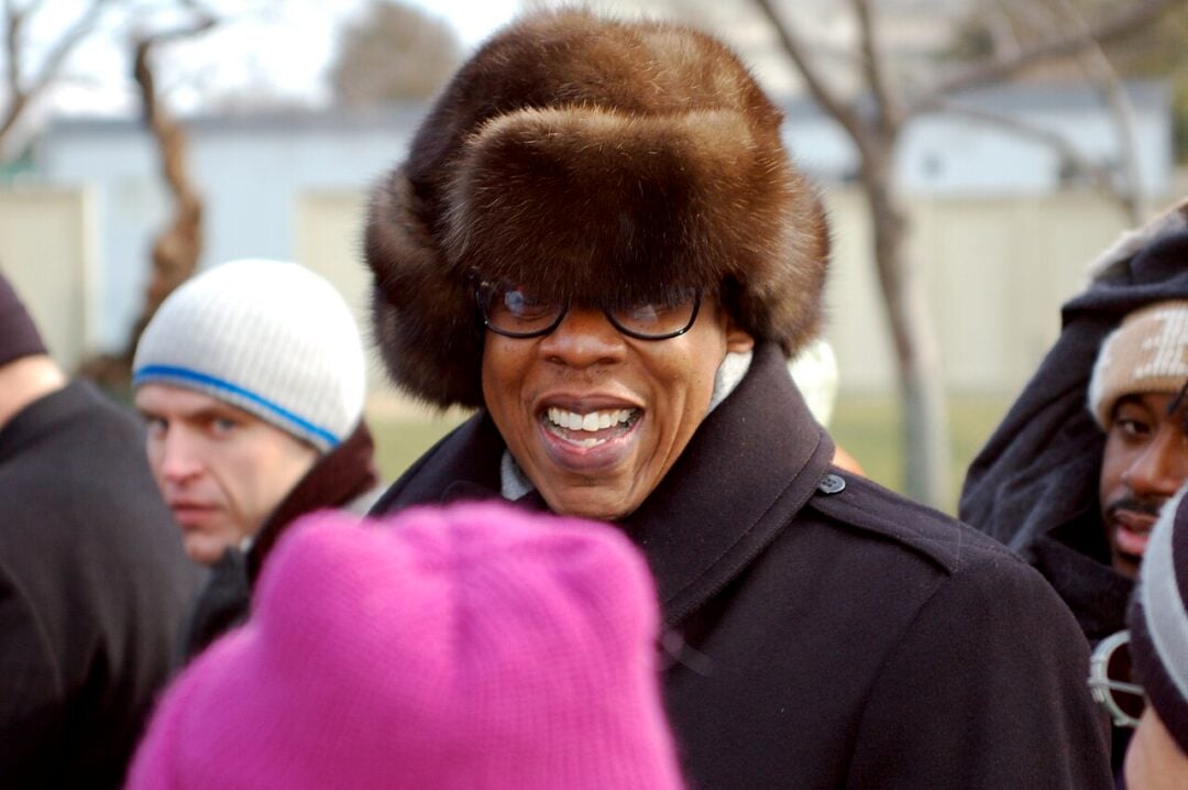A smiling person wearing glasses and a large fur hat stands outdoors among others dressed in winter clothing, including beanies and coats. Trees and a fence are visible in the background.