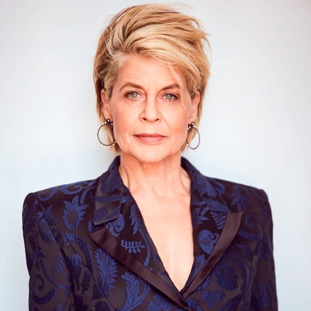A woman with short, blonde hair and hoop earrings wears a dark blue patterned blazer with black lapels. She stands against a light background, looking confidently at the camera.