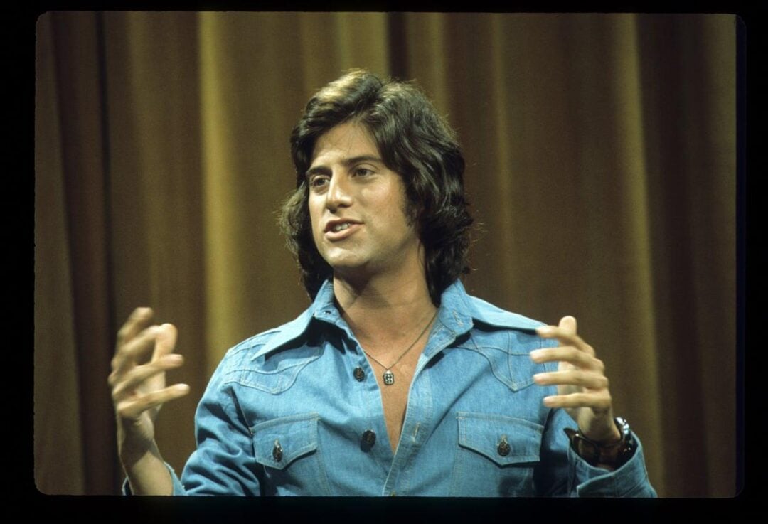 A man with long dark hair wearing a blue denim shirt gestures with both hands while speaking in front of a brown curtain background.