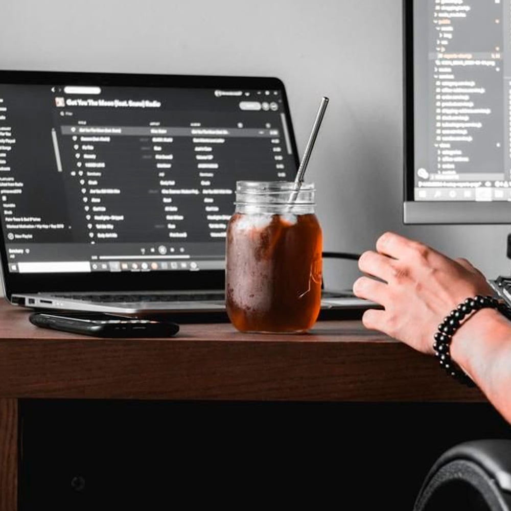 A person’s hand rests on a desk beside an iced drink in a mason jar with a metal straw. A laptop with a music playlist and a monitor displaying code are in the background.