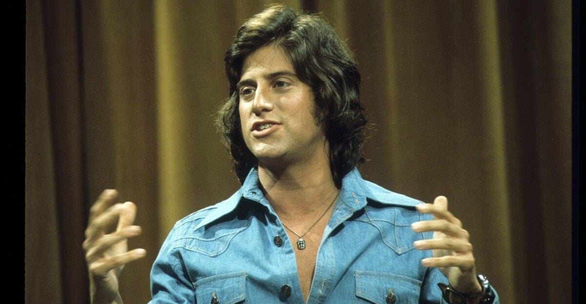 A man with medium-length dark hair wearing a blue button-up shirt gestures with his hands while speaking, standing in front of a brown curtain background.