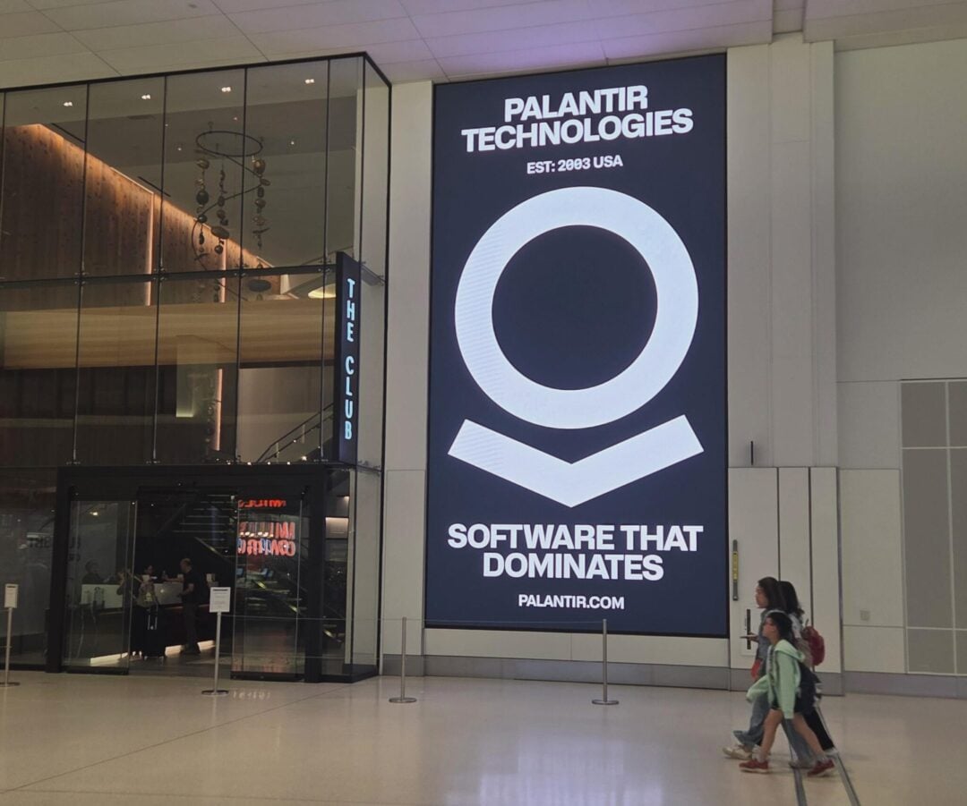 A large digital display in an airport shows "Palantir Technologies" with the text "Software That Dominates" and the website palantir.com. Three people walk by the sign. A lounge called "The Club" is to the left.