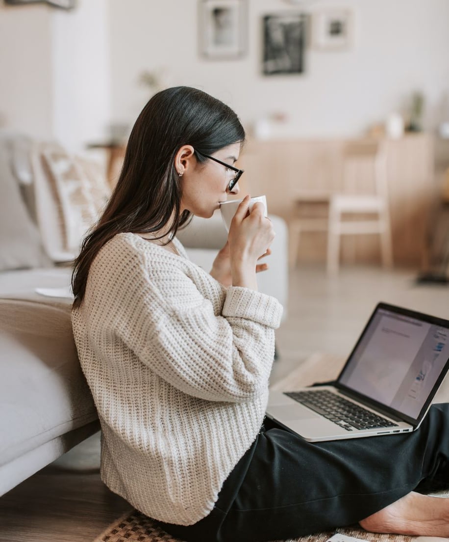 A woman with long dark hair and glasses sits on the floor at home, drinking from a mug and looking at an open laptop. She wears a cozy beige sweater and black pants. The background is softly decorated and blurred.
