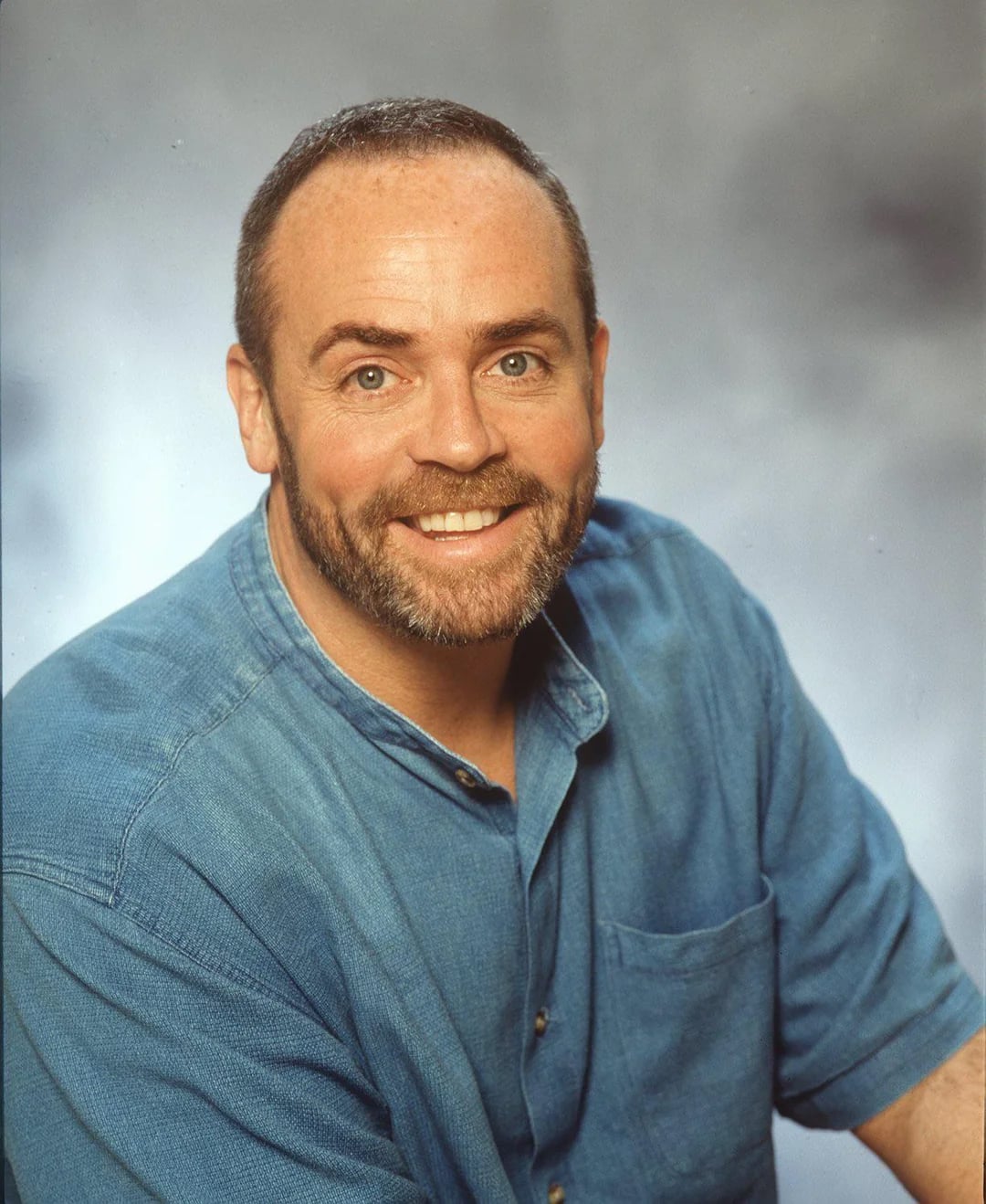 A man with short brown hair, a trimmed beard, and blue eyes is smiling at the camera. He is wearing a blue button-up shirt and is posed against a softly blurred grey background.
