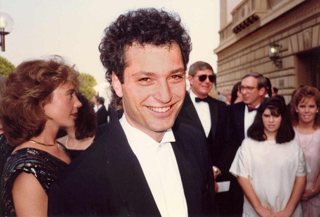 A man in a tuxedo smiles at the camera at an outdoor formal event, surrounded by other people dressed in formal attire. The background shows a building and several attendees.