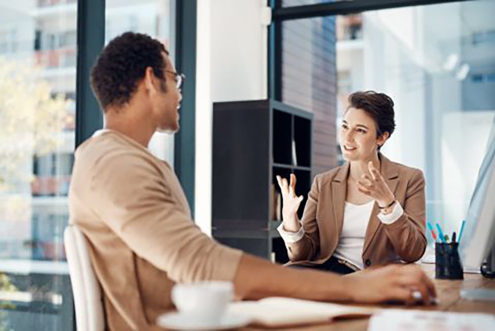 Two people sit across from each other at a desk in a brightly lit office, engaged in a conversation. One gestures with her hands while the other listens attentively, with a computer and coffee cup on the table.