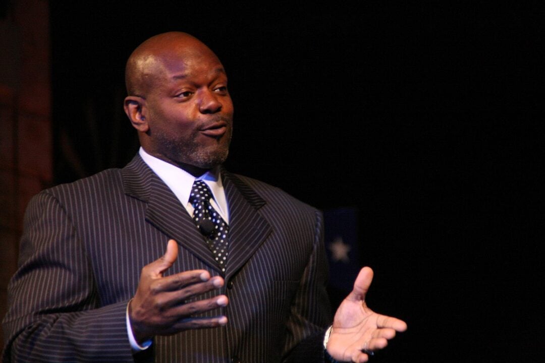 A man in a dark pinstripe suit and polka dot tie gestures with his hands while speaking, set against a dark background.