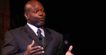 A man in a pinstripe suit and polka dot tie speaks on stage, gesturing with his hands. The background is dark.