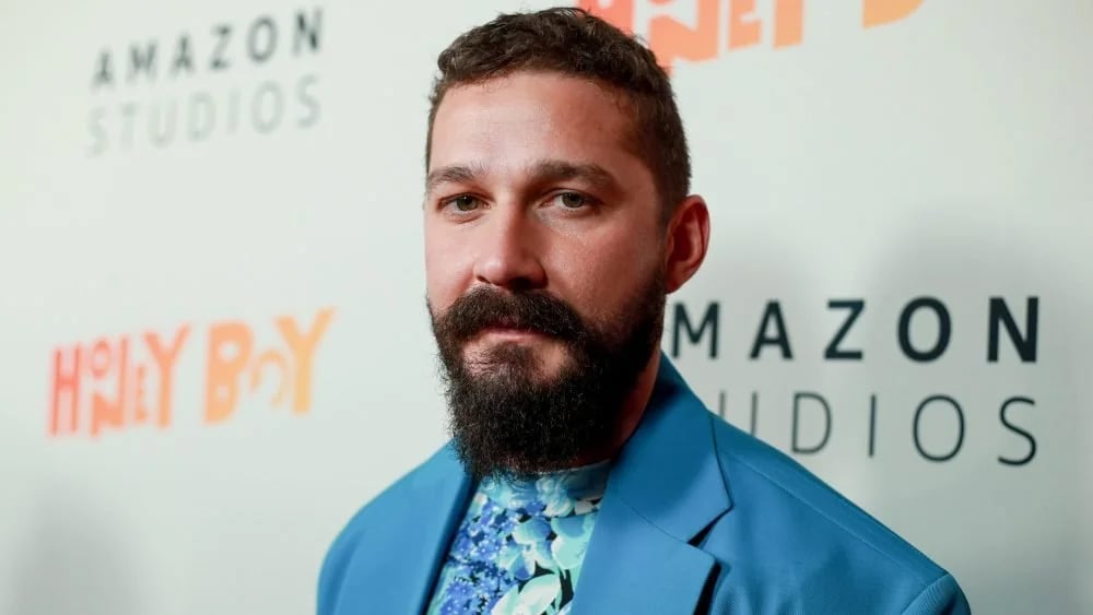 A man with a dark, full beard and short hair wears a blue floral shirt and blue blazer at an event, standing in front of a backdrop displaying "Amazon Studios" and "Honey Boy" logos.