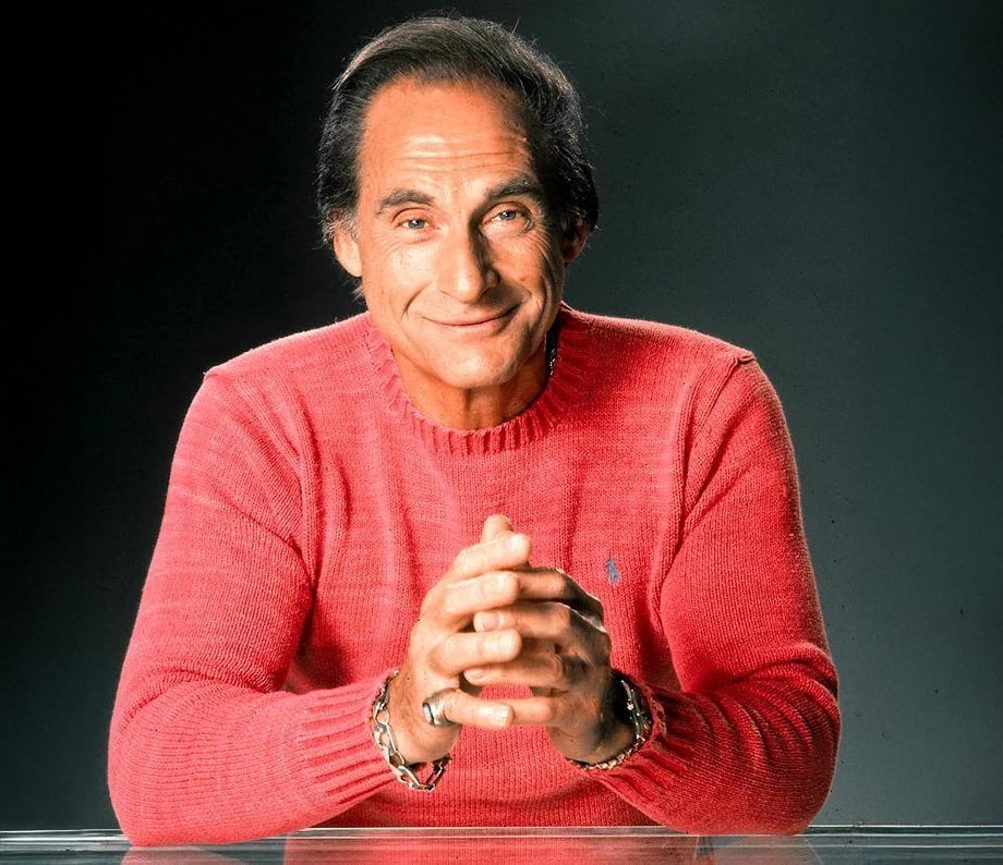 A middle-aged man with dark hair, wearing a bright pink sweater, sits at a clear table with his hands clasped and smiles warmly at the camera against a dark background.