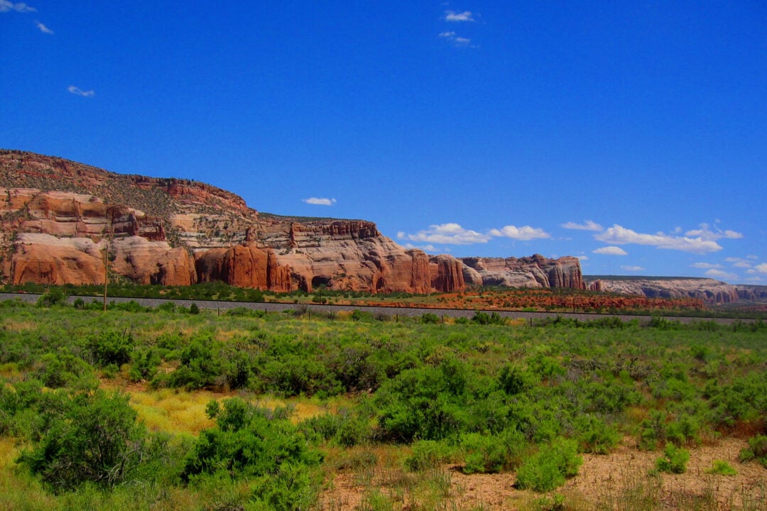 A bright landscape featuring green shrubs in the foreground, a row of reddish-orange rocky cliffs in the distance, and a clear blue sky with a few small clouds overhead.