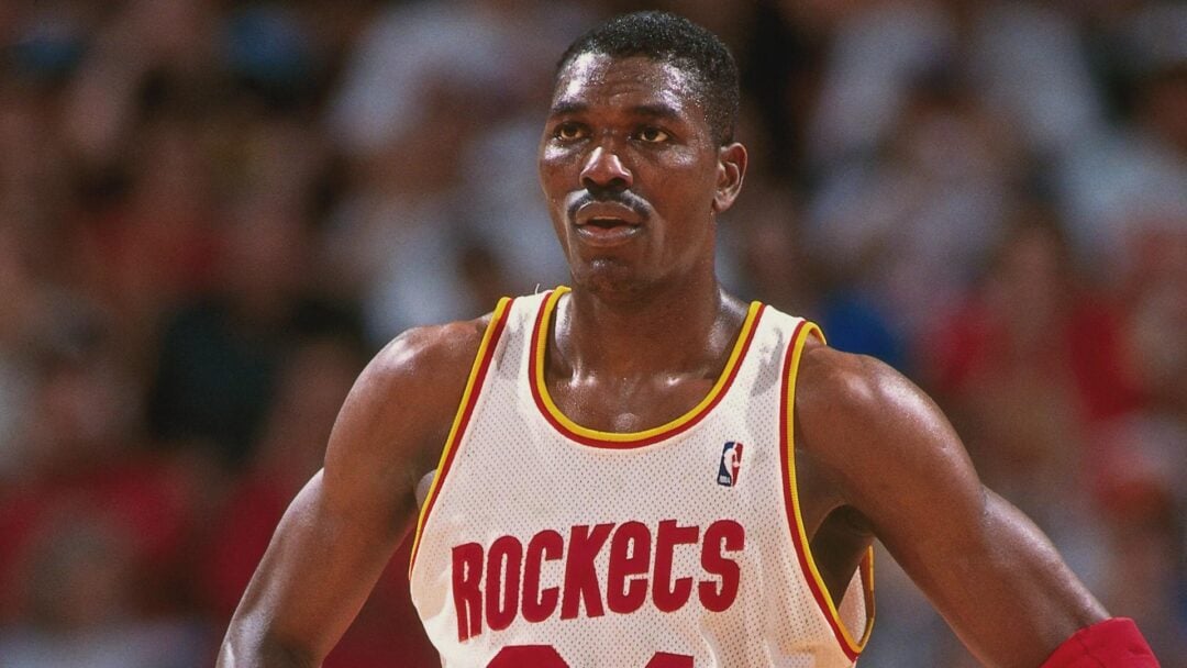 A basketball player wearing a white Houston Rockets jersey with red and yellow trim stands on the court, looking focused. The background shows a blurred crowd.