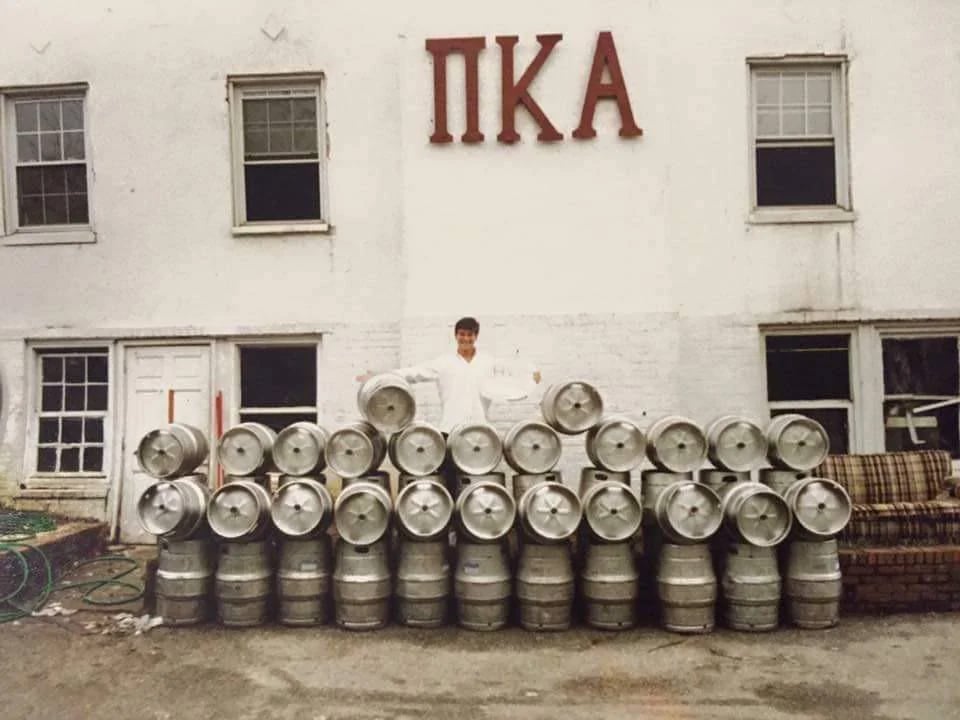A person in white stands behind a large stack of beer kegs arranged in front of a white building with "ΠΚΑ" (Pi Kappa Alpha) displayed above. Four windows and a door are visible on the building.