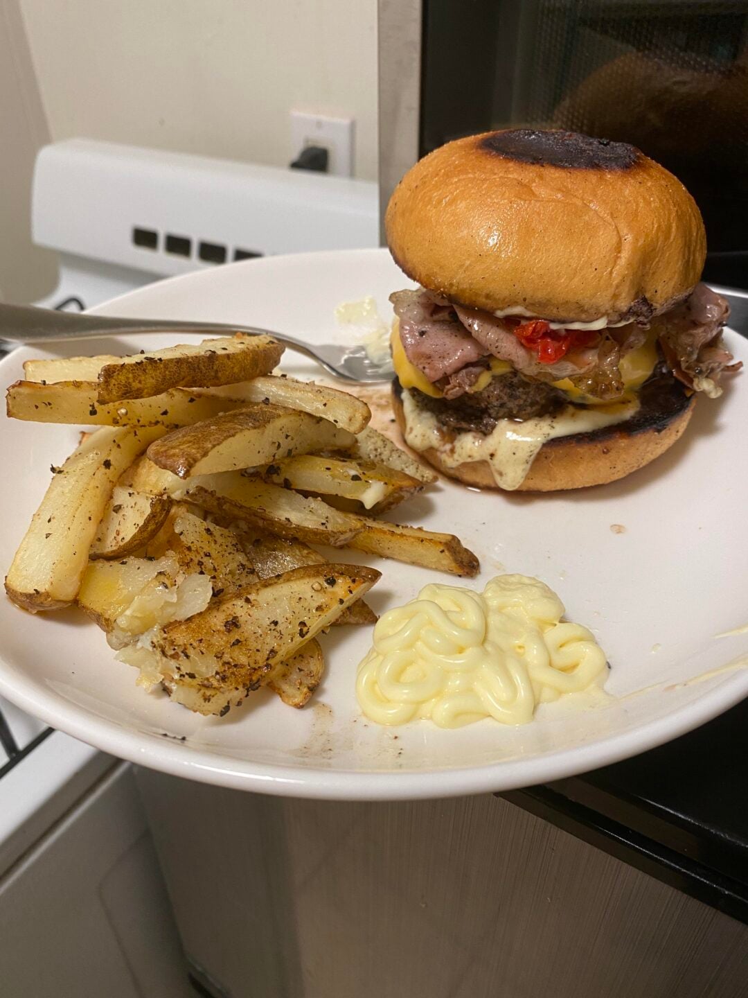 A plate with seasoned potato wedges, a dollop of mayonnaise, and a cheeseburger with roast beef, cheese, and red peppers on a toasted bun, set near a stovetop.