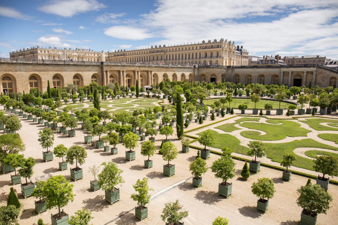 Formal gardens with geometric patterns, trimmed hedges, and potted trees are set in front of the grand, historic Palace of Versailles under a partly cloudy sky.