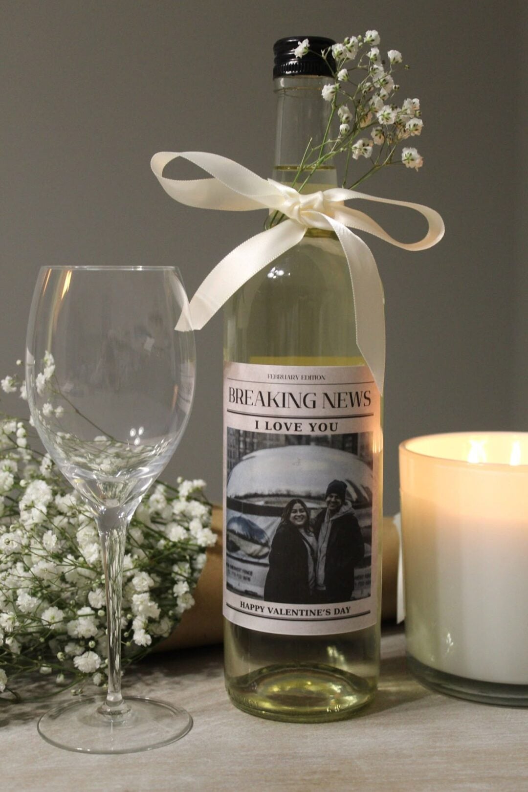 A wine bottle with a white ribbon and flowers, featuring a custom "Breaking News: I Love You" label with a couple’s photo. Next to it are a wine glass, baby's breath flowers, and a lit white candle.