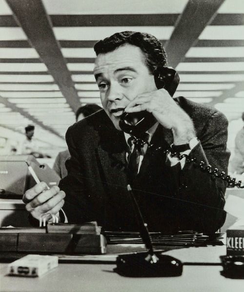 A man in a suit sits at a busy office desk, holding a phone to his ear and another phone in his hand, with papers, a pen, and a box of Kleenex on the desk. The background shows office workers and overhead beams.