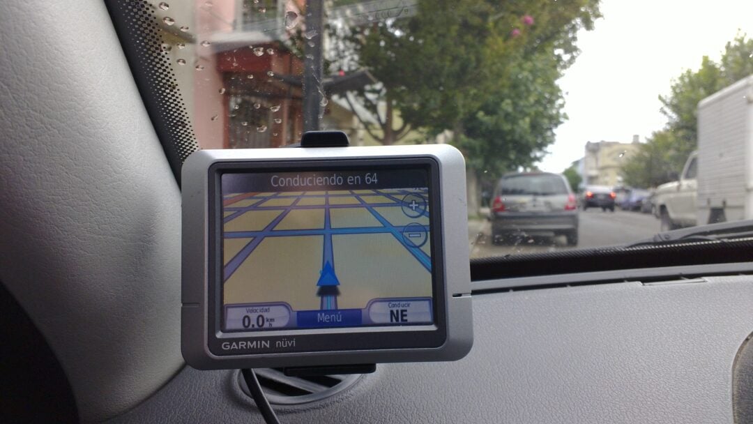 A Garmin nüvi GPS device in a car displays navigation directions in Spanish, showing the speed as 0.0 and the next turn, with a street and parked vehicles visible through the windshield.