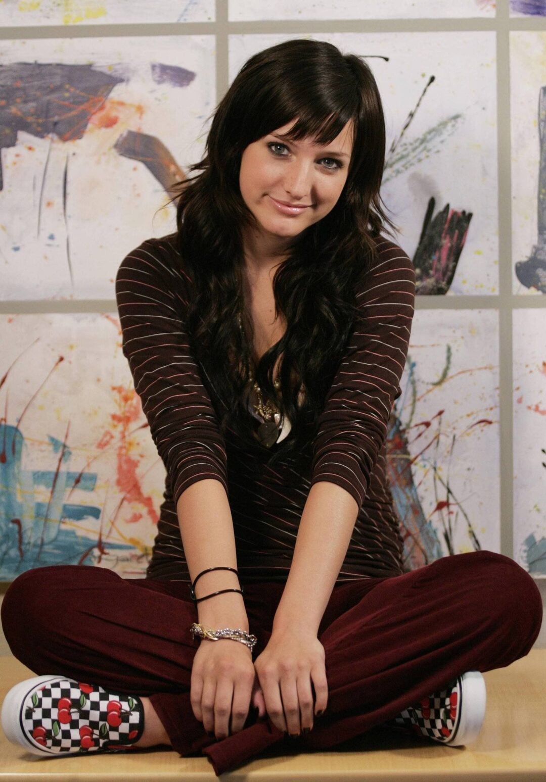 A young woman with long dark hair sits cross-legged on a bench, wearing a striped top, maroon pants, and checkered sneakers. She smiles softly with abstract art visible in the background.